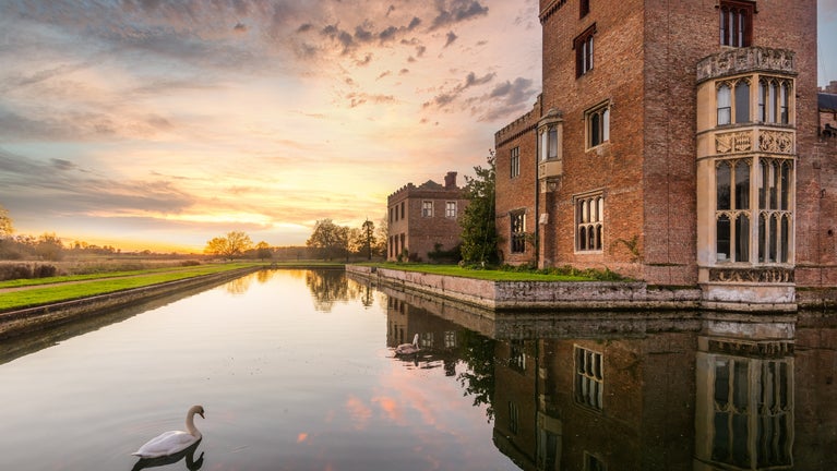 A swan is swimming on the moat surrounding Oxburgh Hall at sunset at Oxburgh Estate.